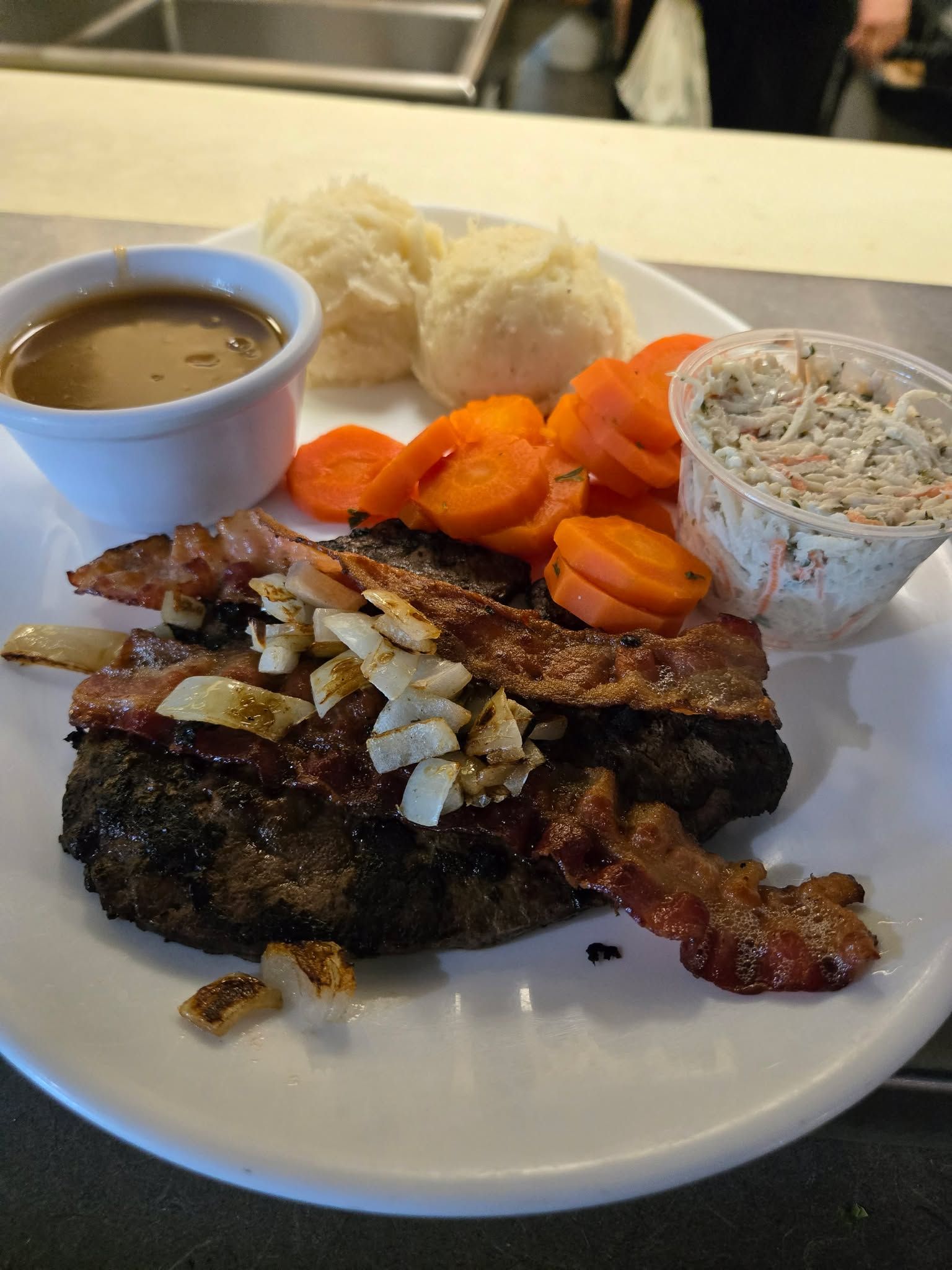 Une assiette composée d'un steak haché garni de bacon, de purée de pommes de terre avec de la sauce, de carottes en rondelles et d'une salade de chou en accompagnement.