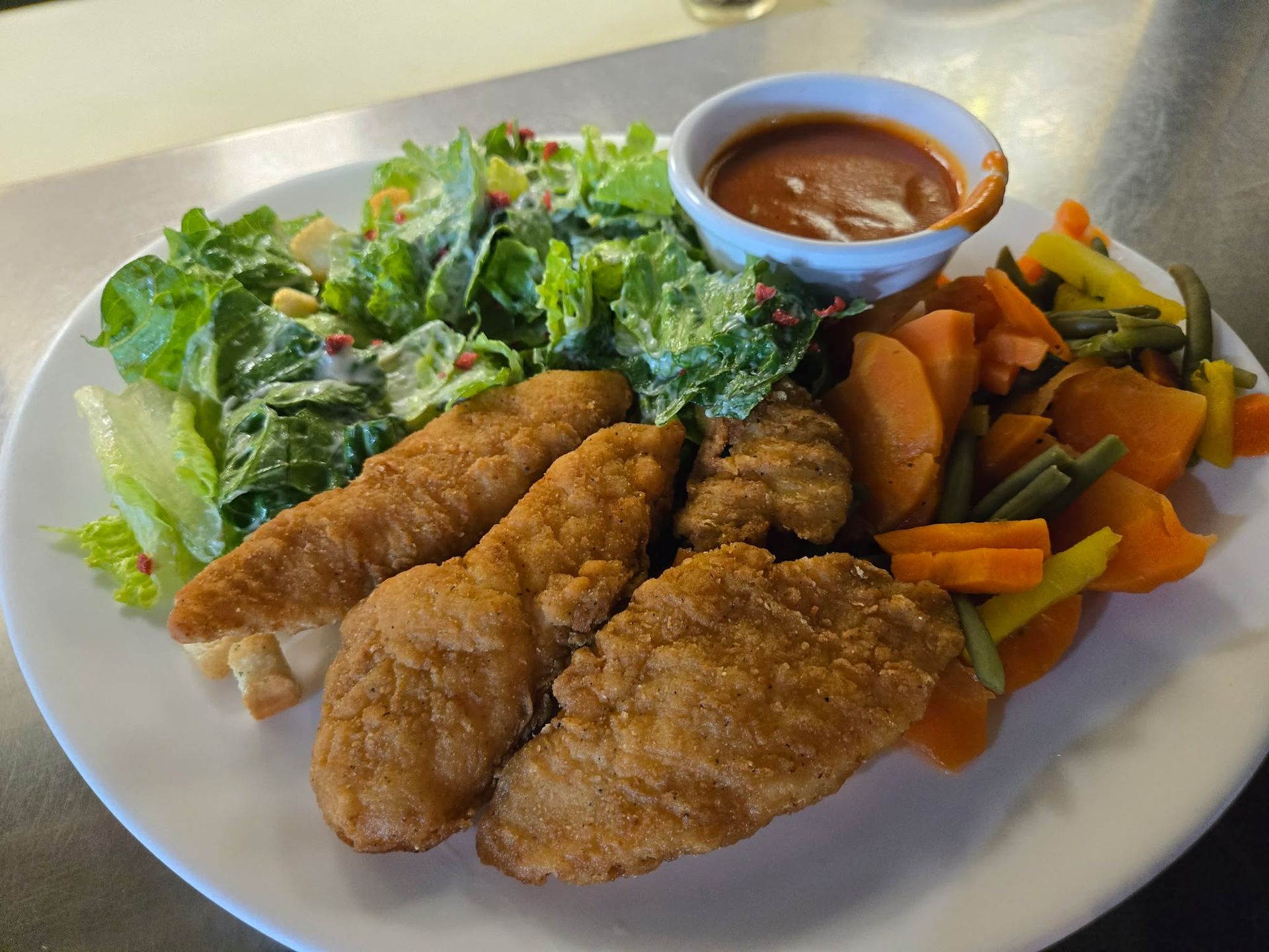 Une assiette composée de filets de poulet panés, d'une salade verte, de carottes et de haricots verts cuits à la vapeur, et d'une sauce à tremper.