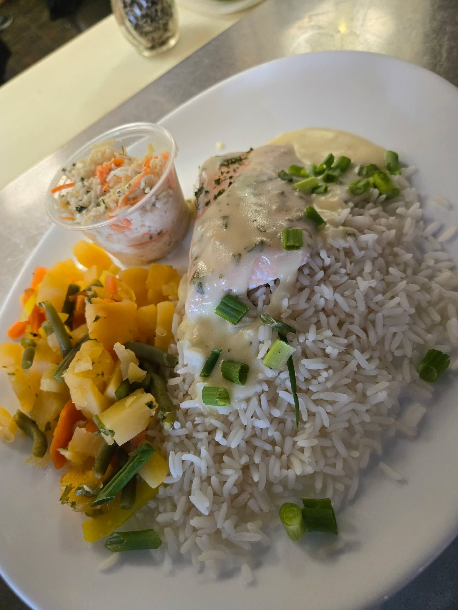 Un plat composé de poisson blanc nappé d'une sauce crémeuse et d'oignons verts, servi avec du riz blanc, des pommes de terre et de la salade de chou.