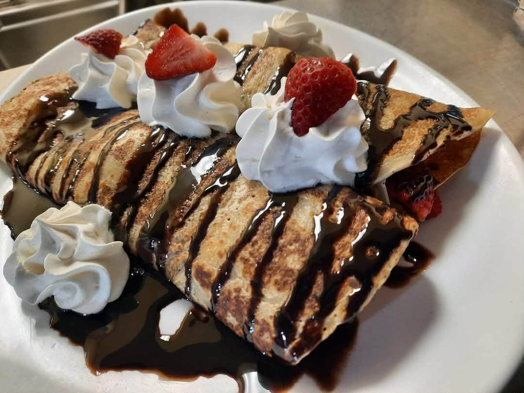 Deux crêpes pliées sur une assiette blanche, arrosées de sauce au chocolat, garnies de crème fouettée et de fraises fraîches.