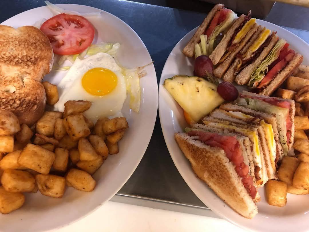 Deux assiettes : l'une avec un œuf au plat, des toasts et des pommes de terre, l'autre avec des club sandwiches, des fruits et des pommes de terre.