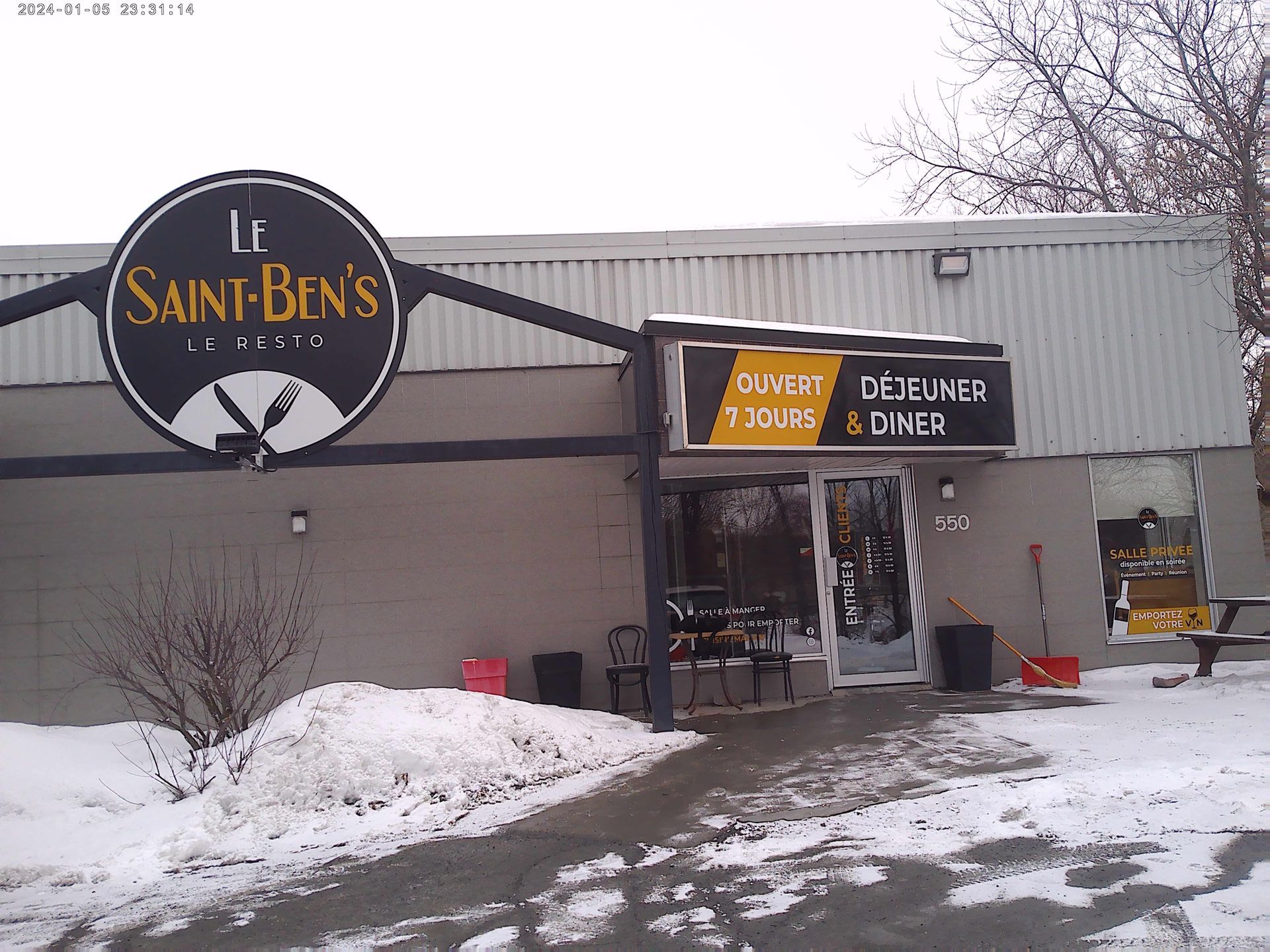 L'entrée d'un restaurant nommé Le Saint-Ben's, avec une enseigne circulaire noire et des menus jaunes sur fond enneigé.