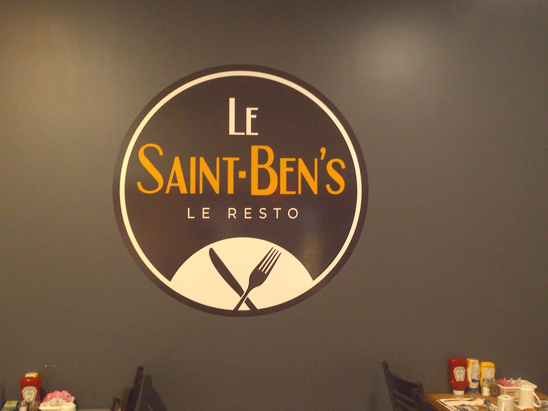 Une enseigne circulaire pour « Le Saint-Ben's Le Resto » avec un texte doré et une icône blanche représentant un couteau et une fourchette sur un mur sombre.