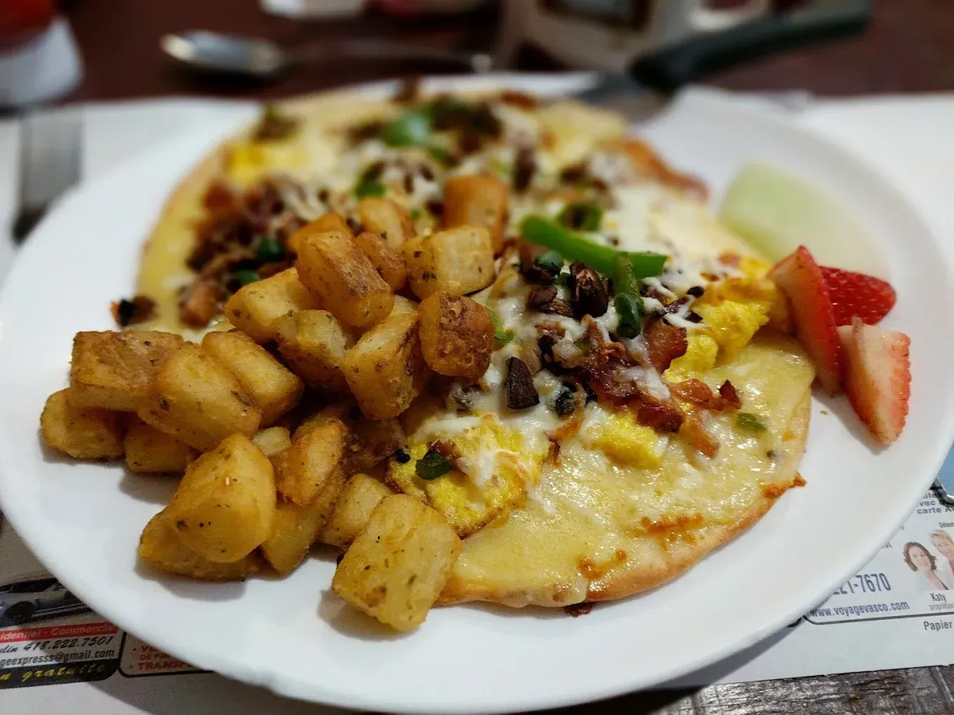 Une assiette de petit-déjeuner composée d'une grande omelette recouverte de fromage fondu, de pommes de terre croustillantes coupées en dés et de deux tranches de fraises.