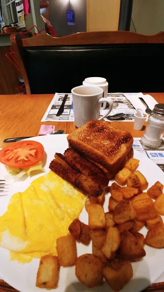 Une assiette de petit-déjeuner composée d'une omelette, de pommes de terre rôties, de deux tranches de pain grillé et d'une tranche de tomate, à côté d'une tasse à café.