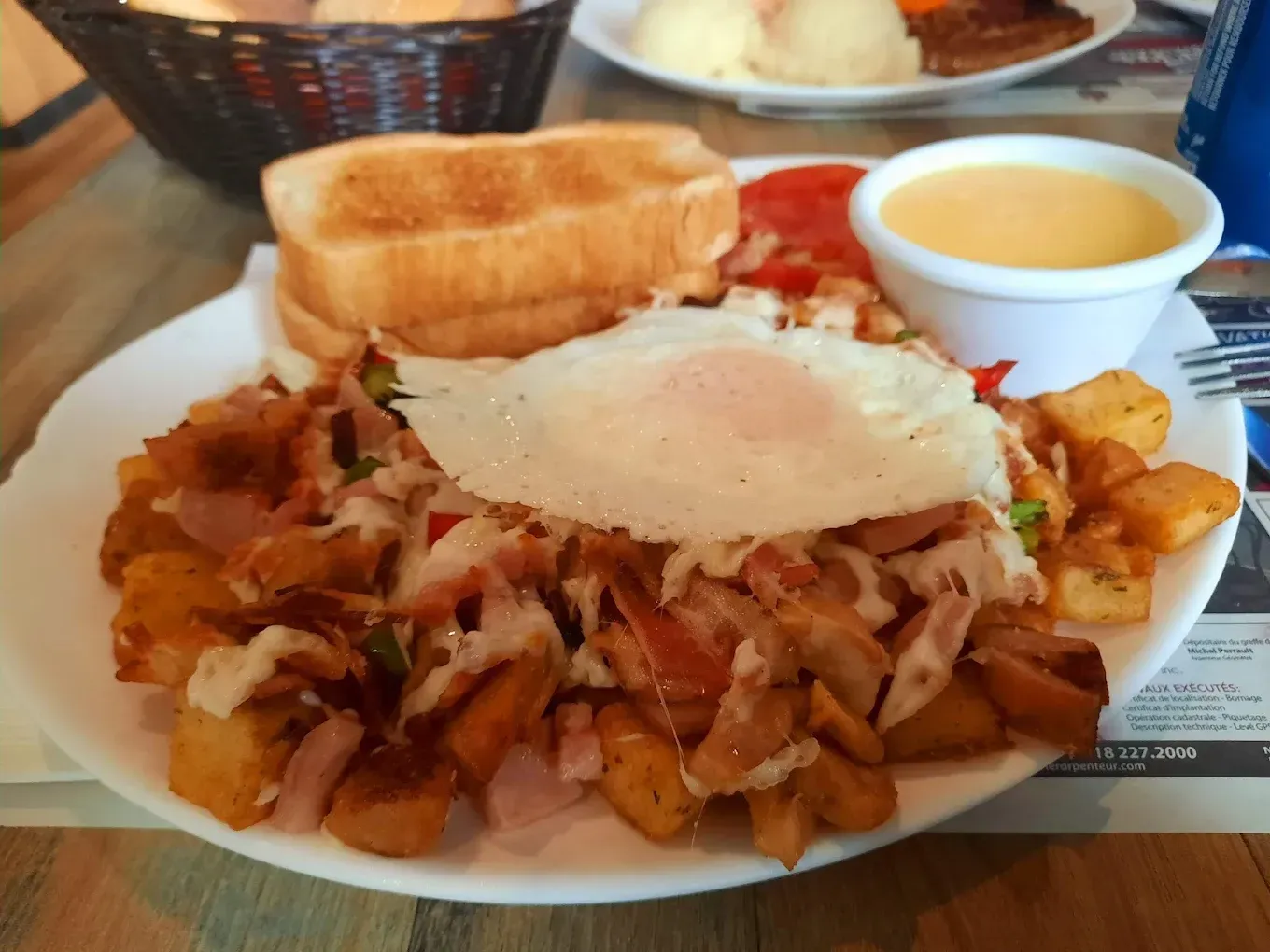 Une assiette de petit-déjeuner composée de pommes de terre en dés avec de la viande, du fromage fondu et un œuf au plat, servie avec des toasts et une coupelle de sauce.