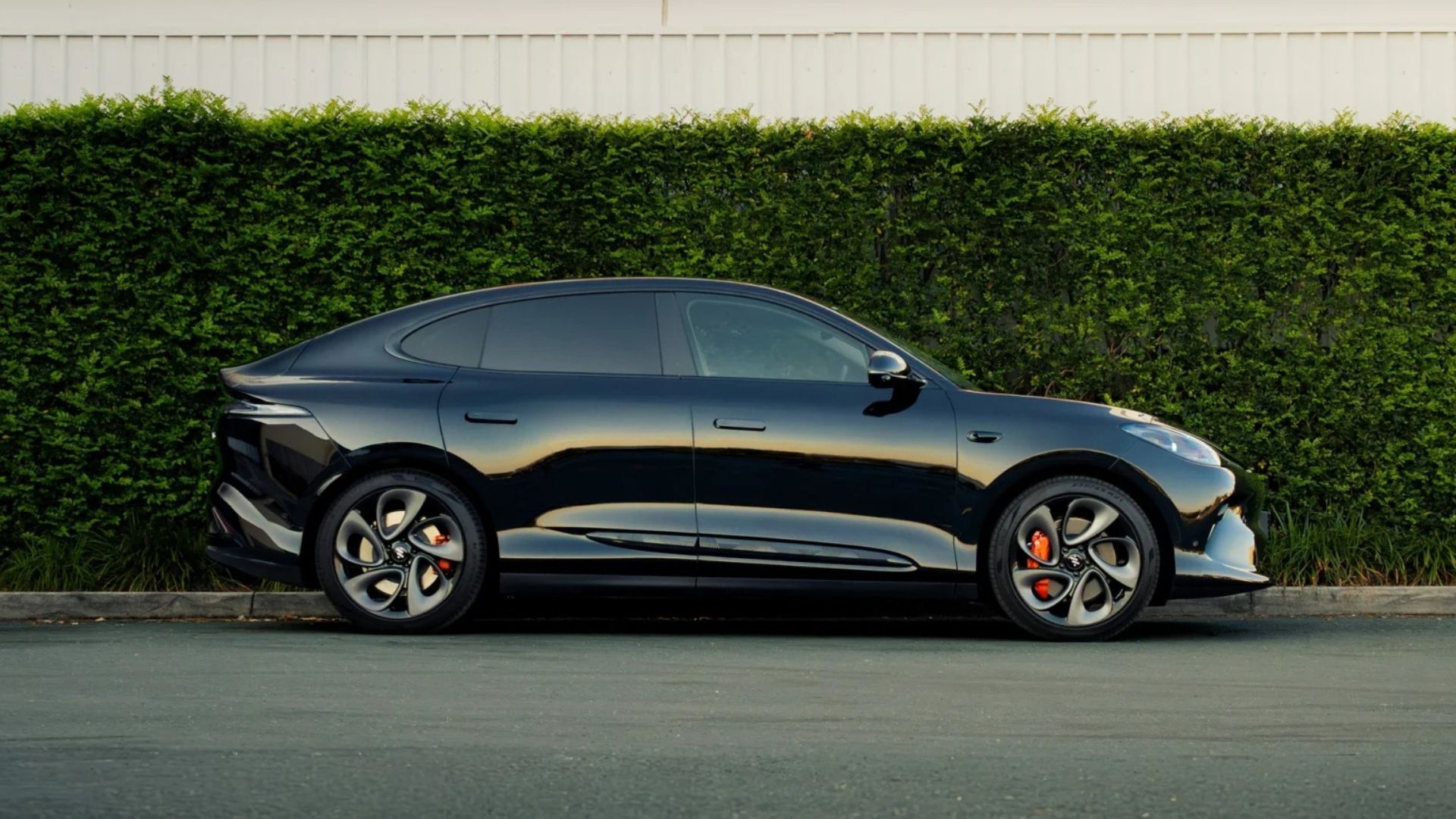 Black sedan parked beside a tall green hedge and beige wall on a street