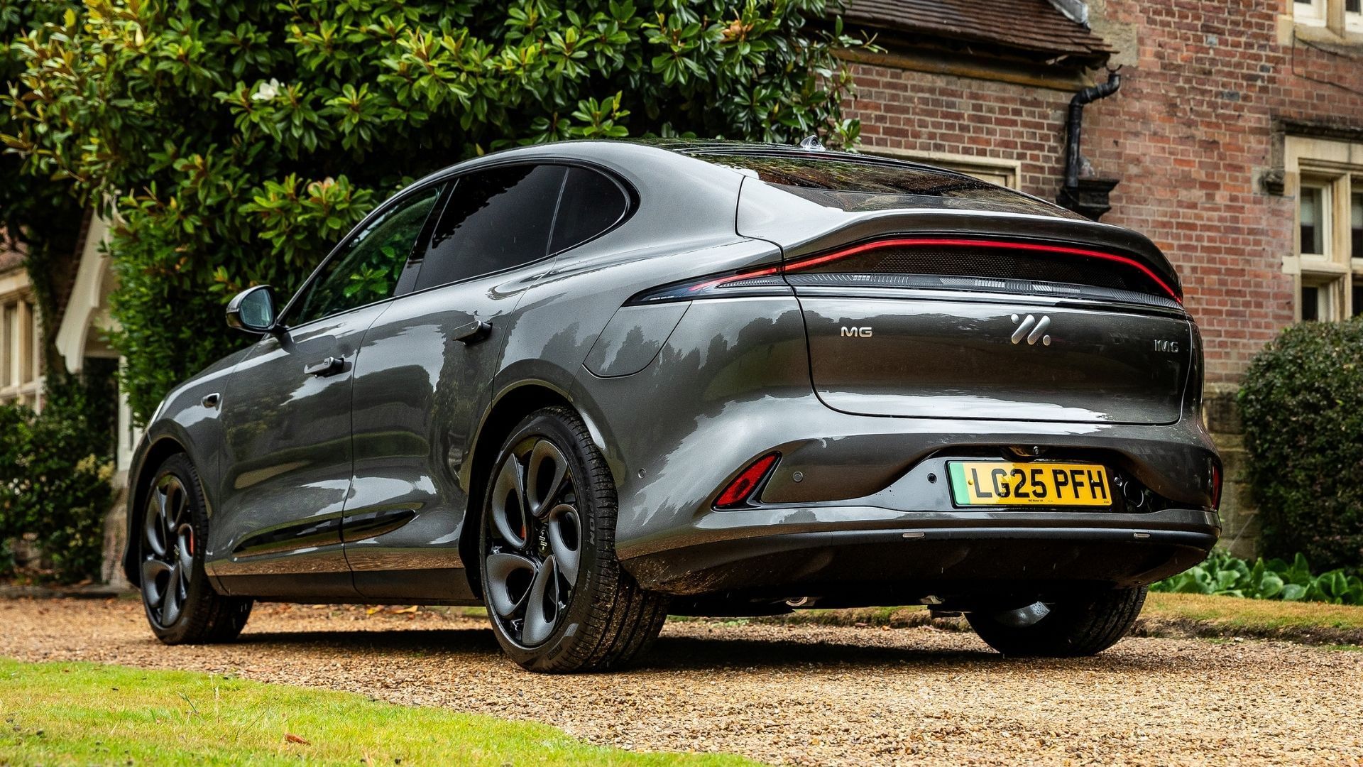 Dark gray sports sedan parked on a gravel driveway beside a brick house and greenery, rear three-quarter view