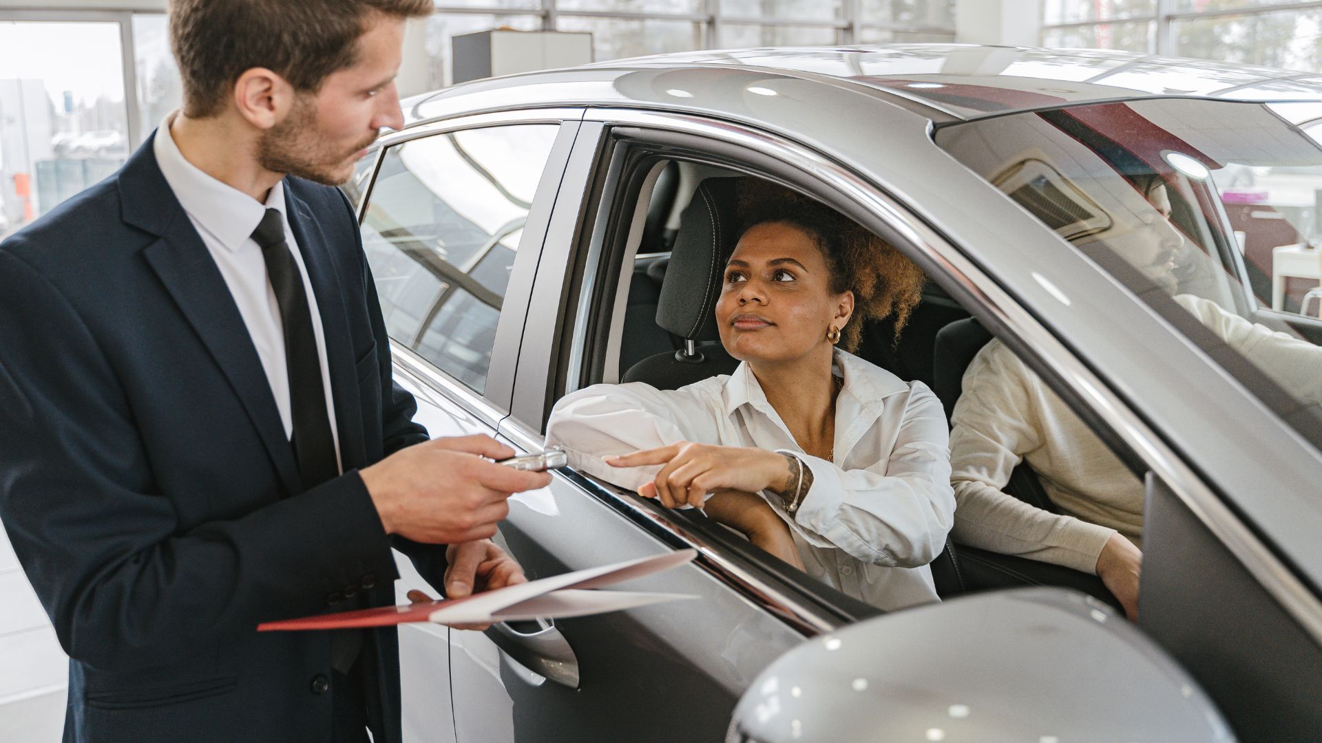 Salesman showing car features to a customer seated inside a silver car at a dealership