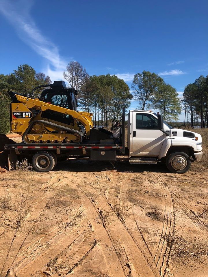 Tow truck carrying a yellow skid steer on a dirt lot under a blue sky
