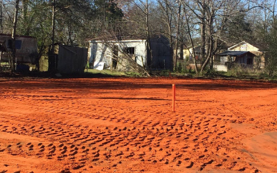 Freshly graded orange dirt lot with a small marker stake, houses and bare trees in the background