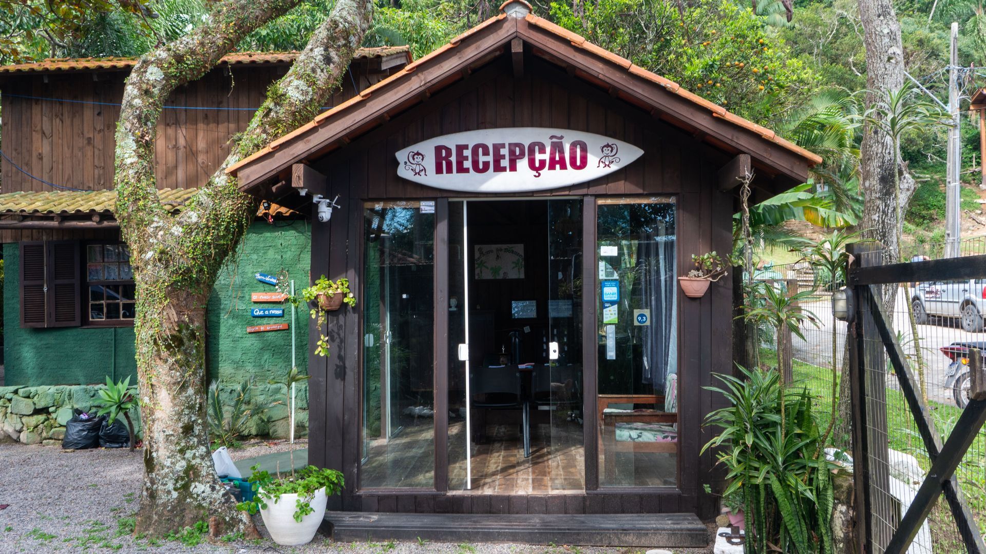 Entrada da recepção de um alojamento rústico, com portas de vidro, placa com a inscrição «RECEPÇÃO» e vegetação exuberante à volta da entrada