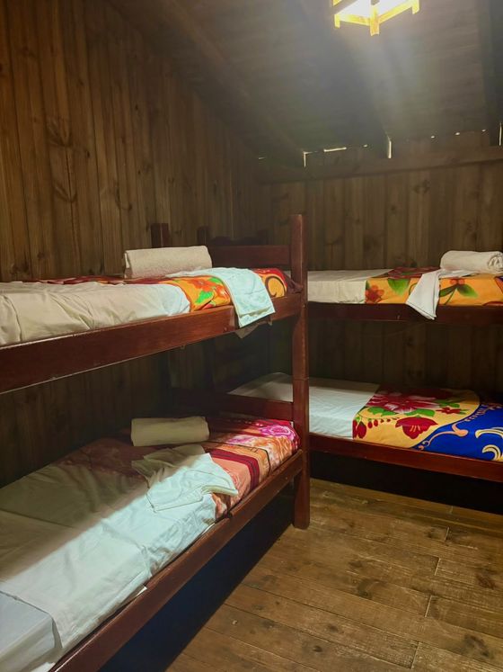 Quarto com beliches revestido de madeira, com dois beliches de madeira, roupa de cama colorida, piso sem revestimento e luminária no teto.