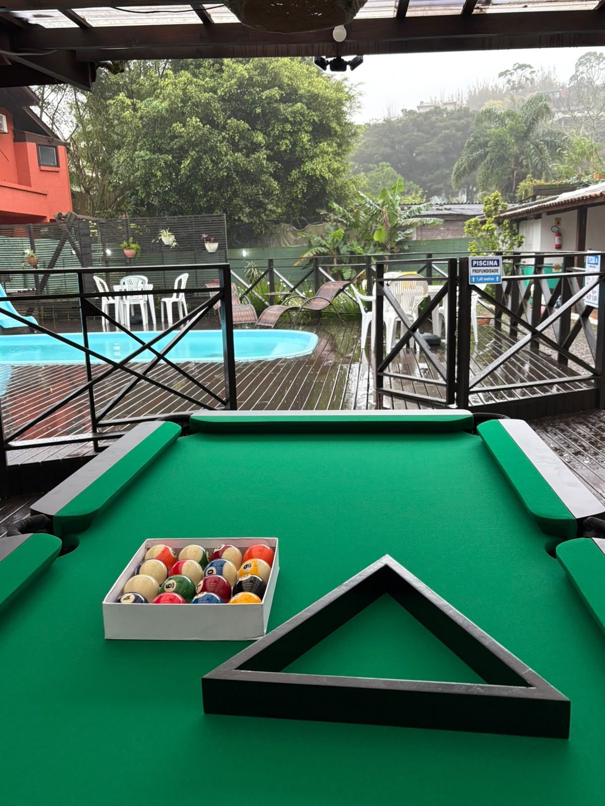 Mesa de bilhar verde com bolas dispostas num triângulo numa varanda chuvosa com vista para uma piscina e árvores