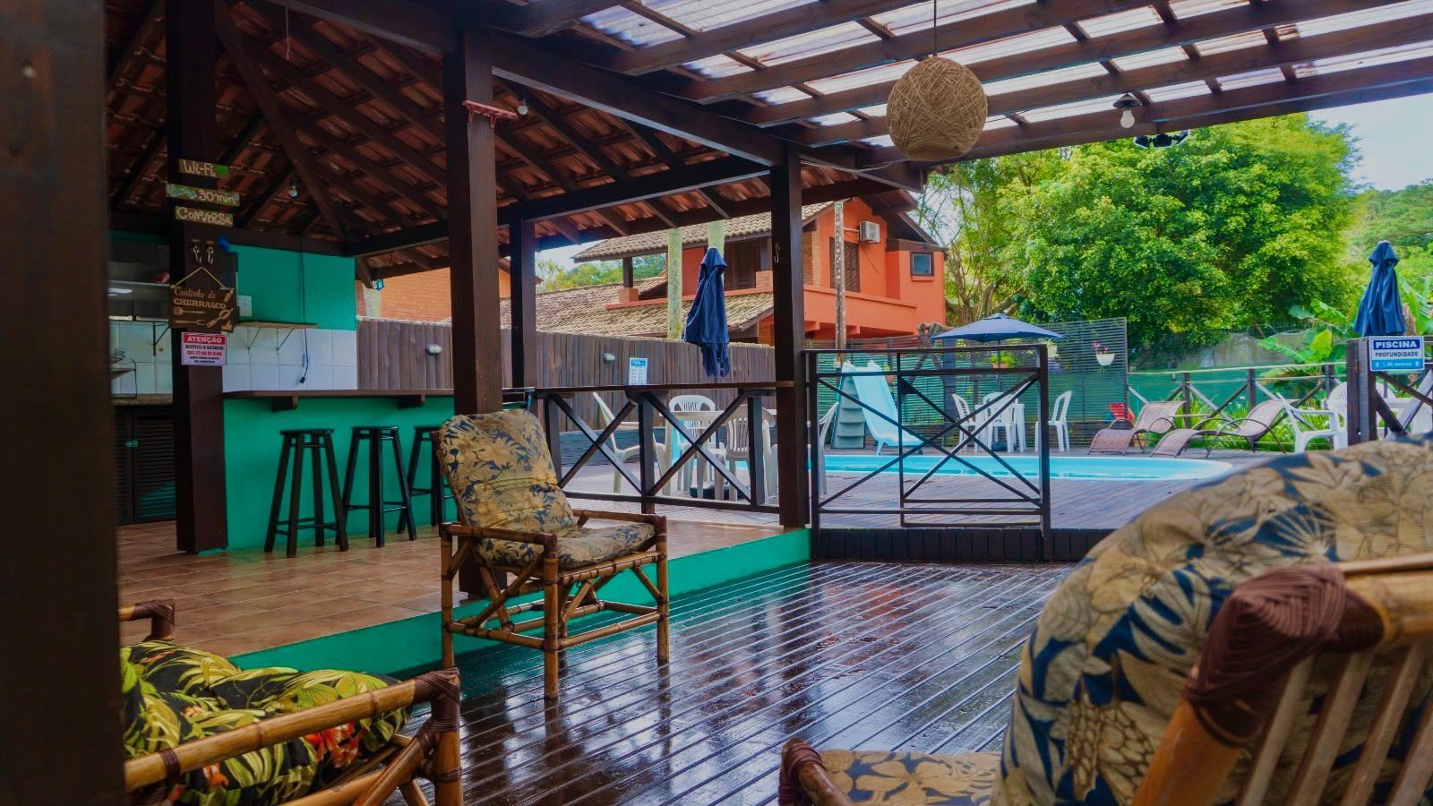 Esplanada coberta de café ao ar livre, com mesas turquesa, piso de tijolos molhado e vista para a piscina após a chuva