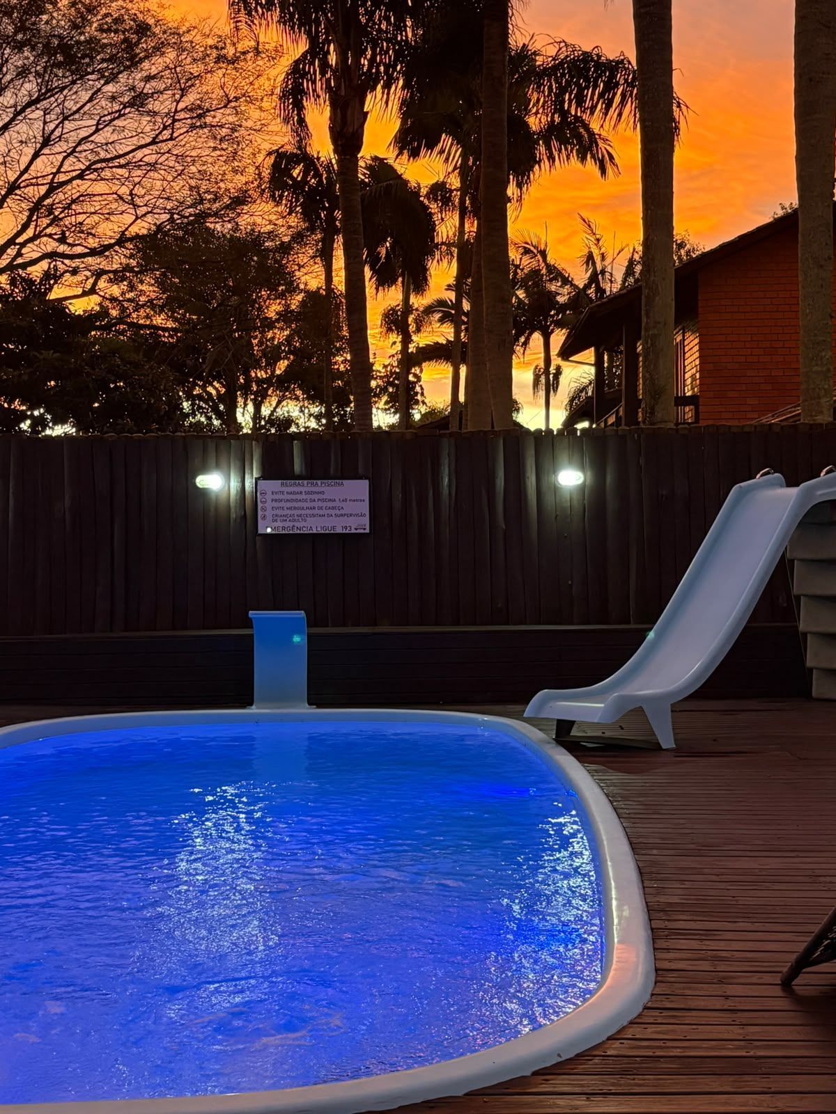 Piscina iluminada em azul e escorrega no quintal ao pôr do sol, com palmeiras e deck de madeira.