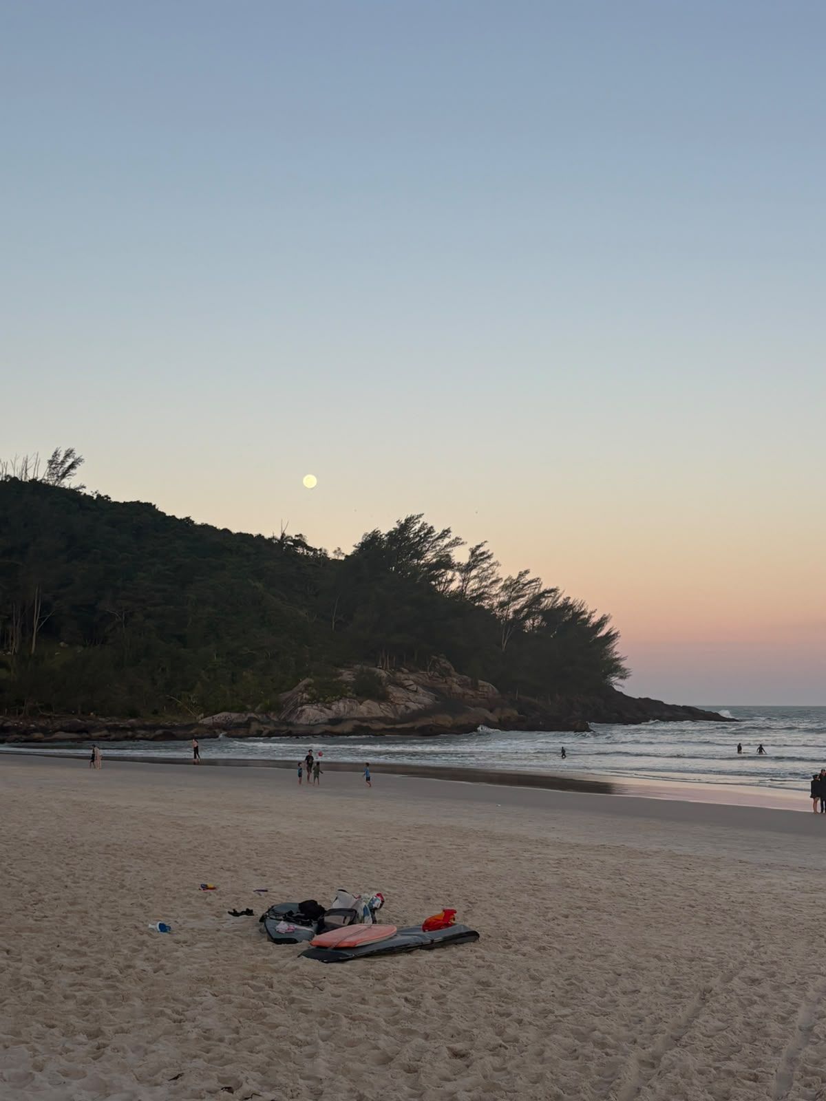 Praia ao pôr do sol, com pessoas caminhando pela orla e pranchas de surfe na areia
