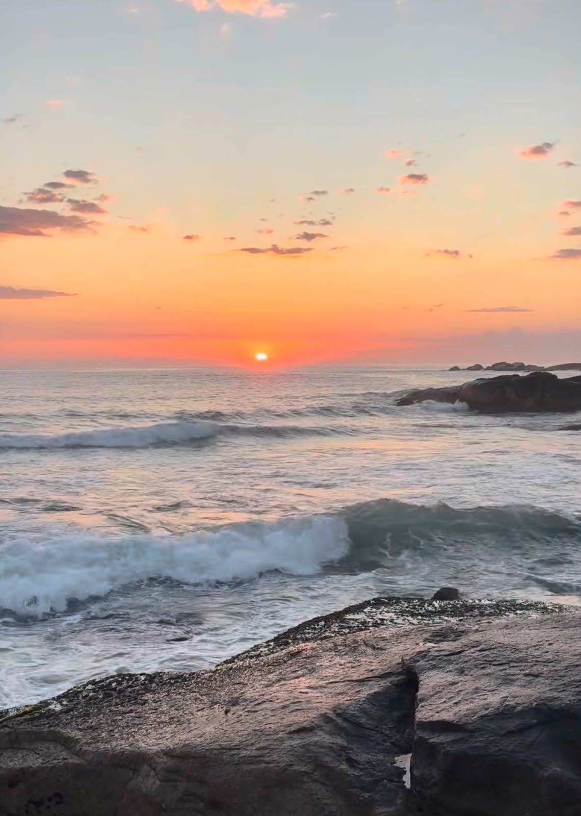 Pôr do sol sobre uma costa rochosa, com as ondas a rebentar e um céu cor-de-rosa e laranja