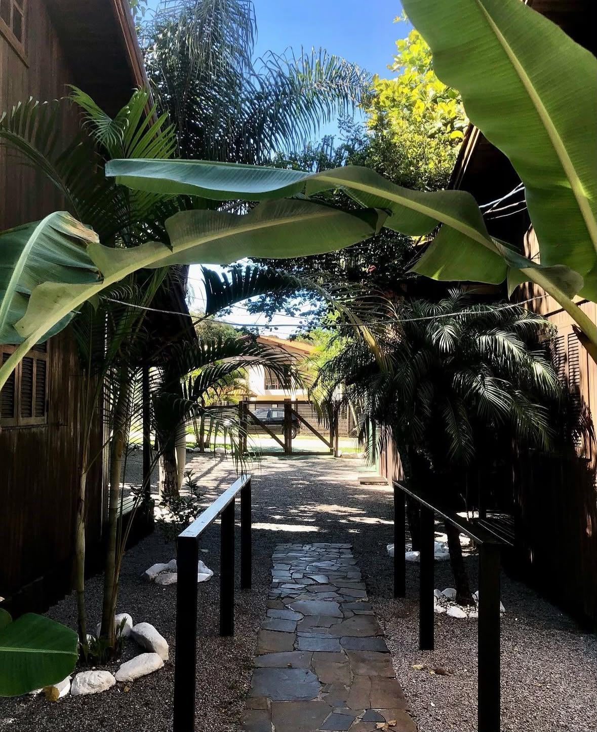 Caminho de pedra por um jardim tropical, com folhas de bananeira formando um arco sobre ele e um gazebo ao longe