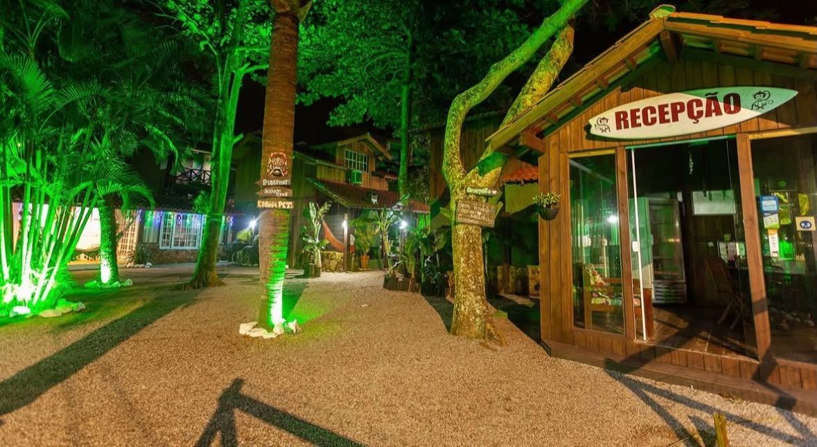 Passarela de resort à noite, com palmeiras, iluminação verde e uma placa indicando a entrada da recepção.