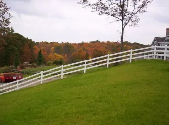 Grassy hillside with a white fence, a red car, and autumn trees under a cloudy sky