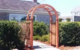 Wooden garden archway with lattice sides over a path, framed by tall hedges beside a house.