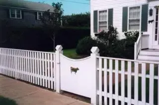 White picket fence with a curved gate and small dog emblem in front of a house and driveway
