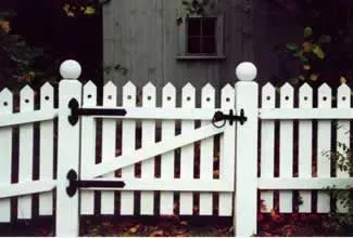 White picket gate with black hinges and latch in front of a dark fence and house