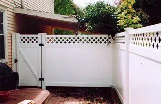 White vinyl fenced backyard patio with a gate and lattice top, beside a house and grill