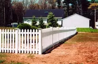 White picket fence along a dirt yard beside a white house and garage with trees in the background
