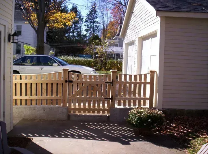 Wooden fence and gate in front of a beige garage and house, with a driveway and parked car nearby.
