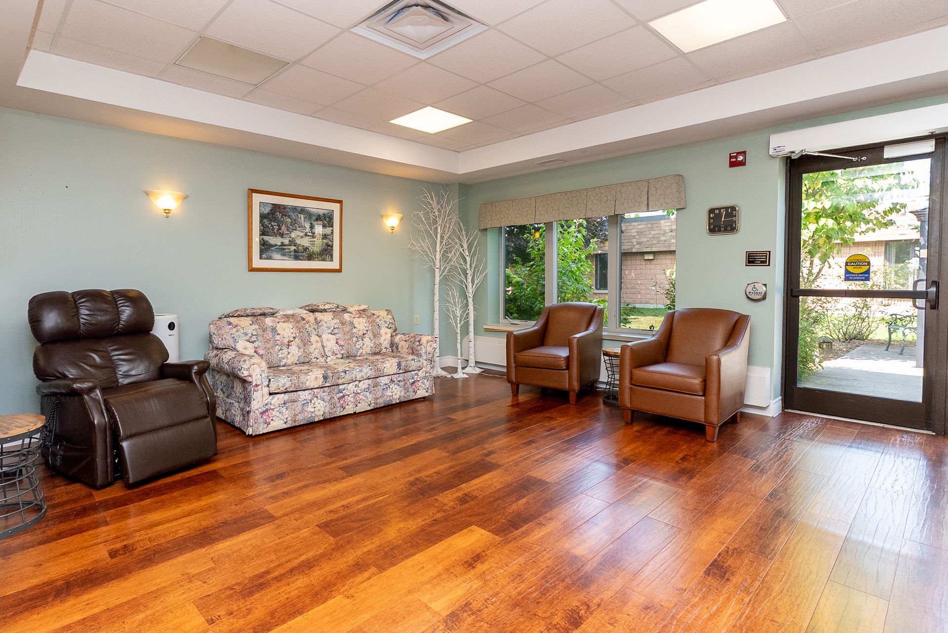 Bright lounge with wood floors, recliners, floral couch, and large windows letting in natural light