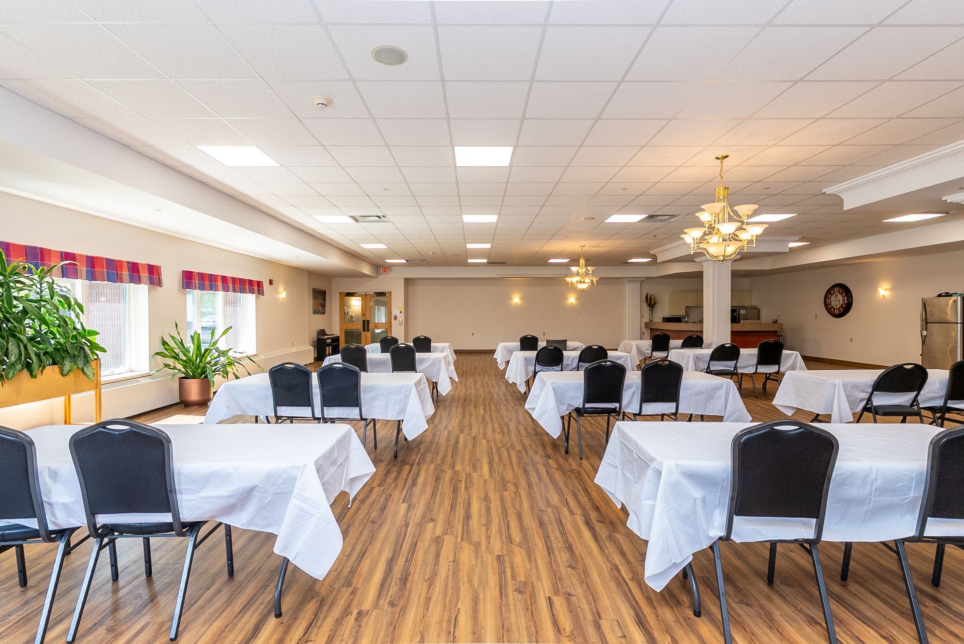 Bright banquet hall with white tablecloths, black chairs, wood floors, and plants by the windows.