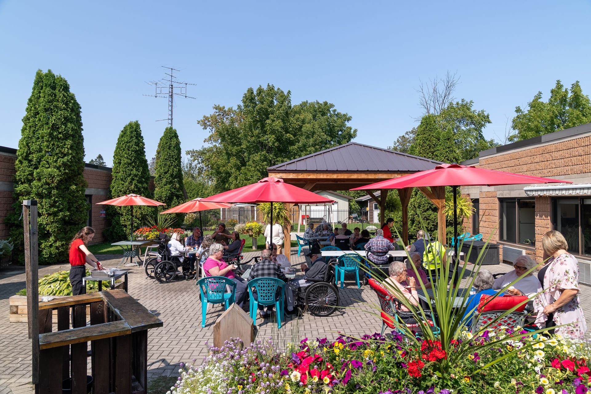 Outdoor café patio with red umbrellas, seated patrons, and colorful flowers in bright sunlight