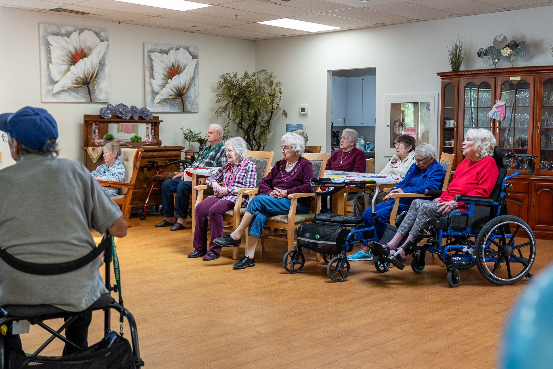 Residents seated in a common room watching a speaker, with wheelchairs and framed wall art nearby