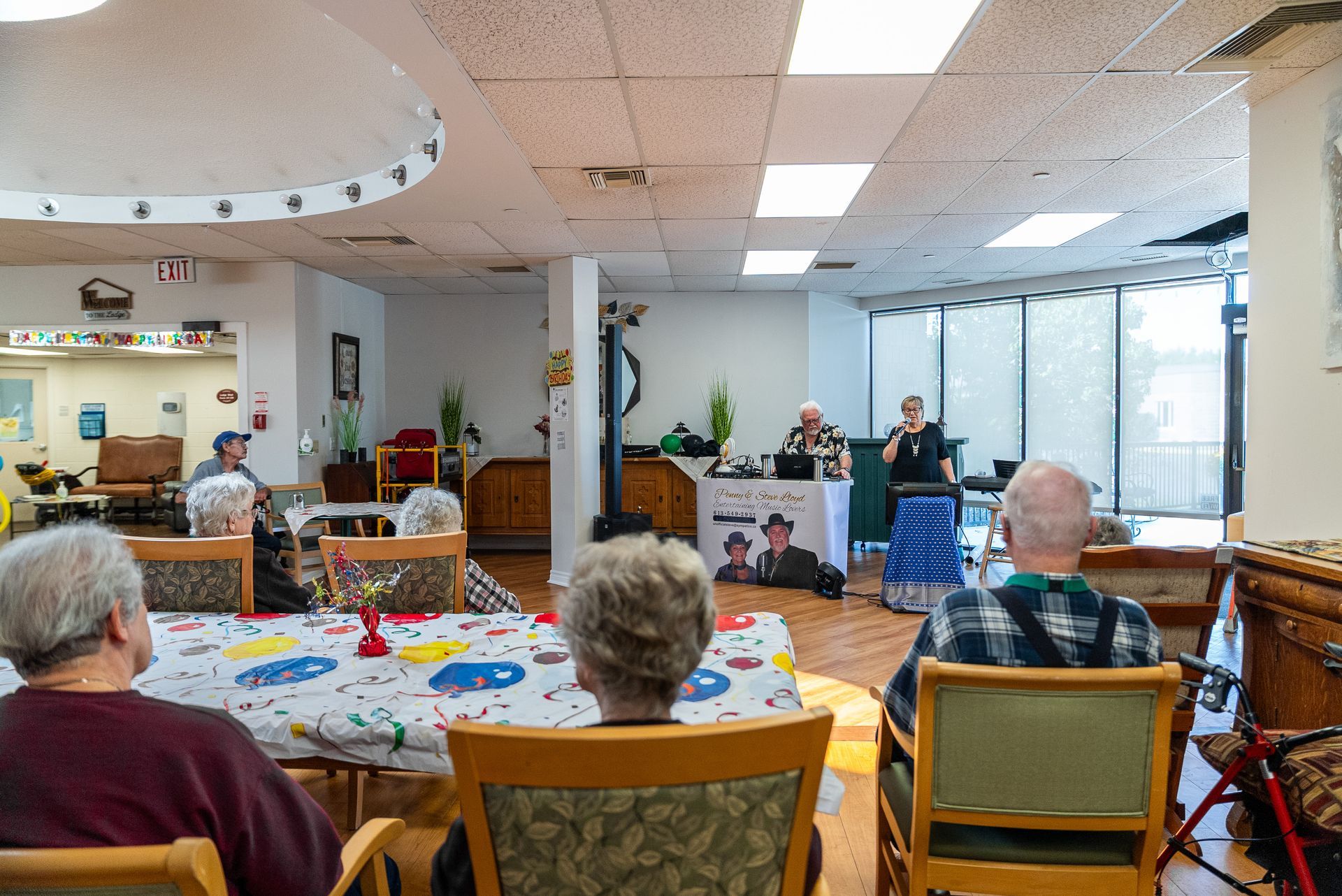Residents sit at tables in a bright community room, watching a small group perform near large windows.
