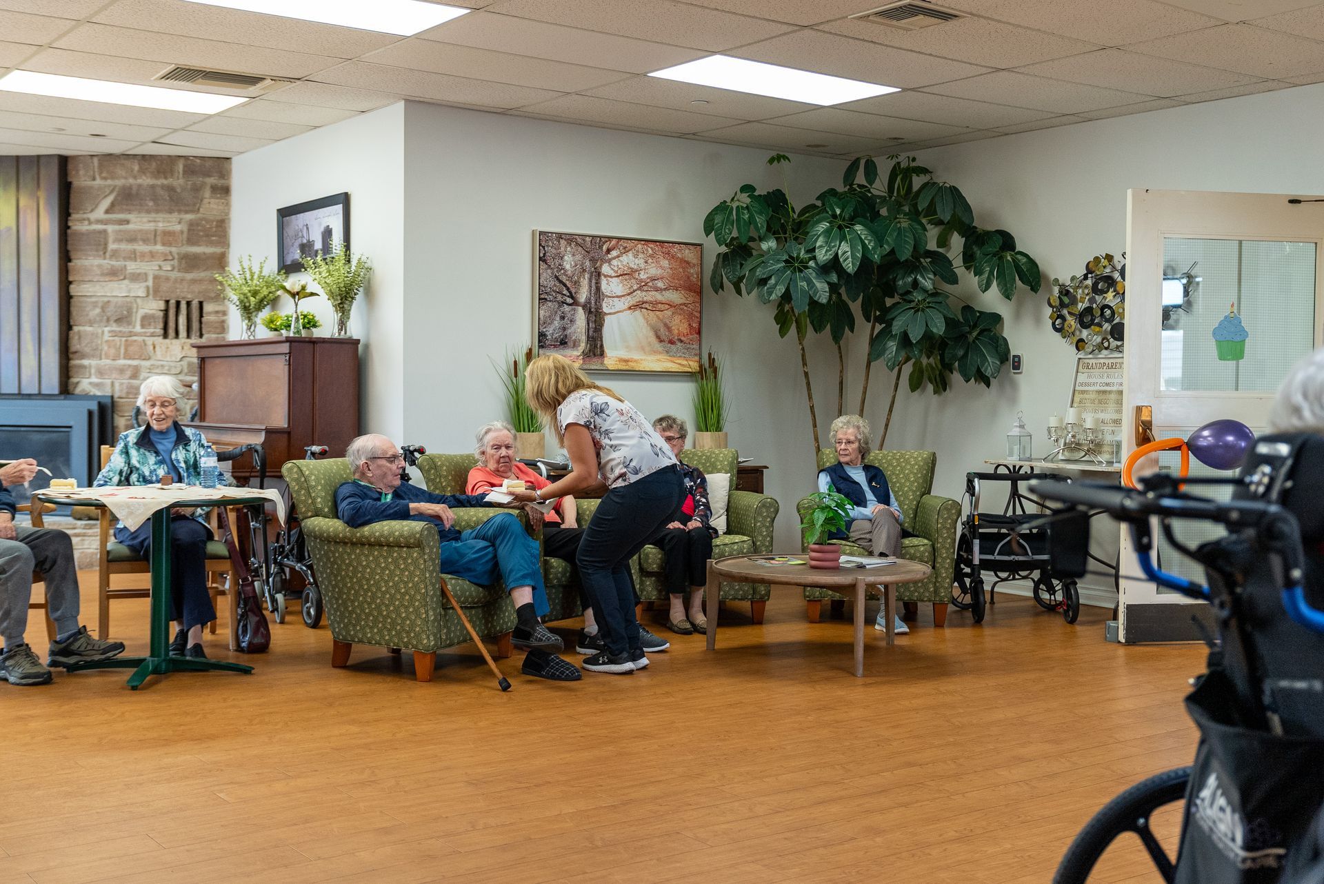 People sitting and chatting in a cozy community room with chairs, tables, plants, and a tiled floor.