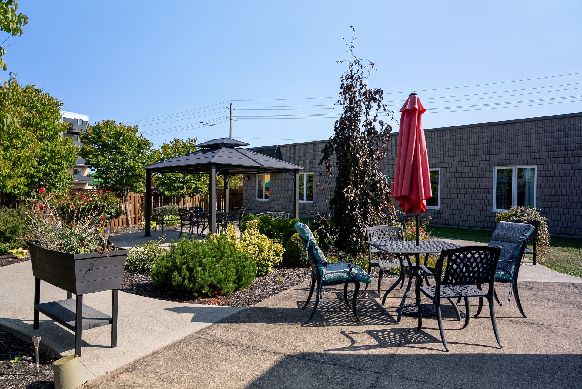 Outdoor patio with tables, chairs, red umbrella, and landscaped garden beside a stone building