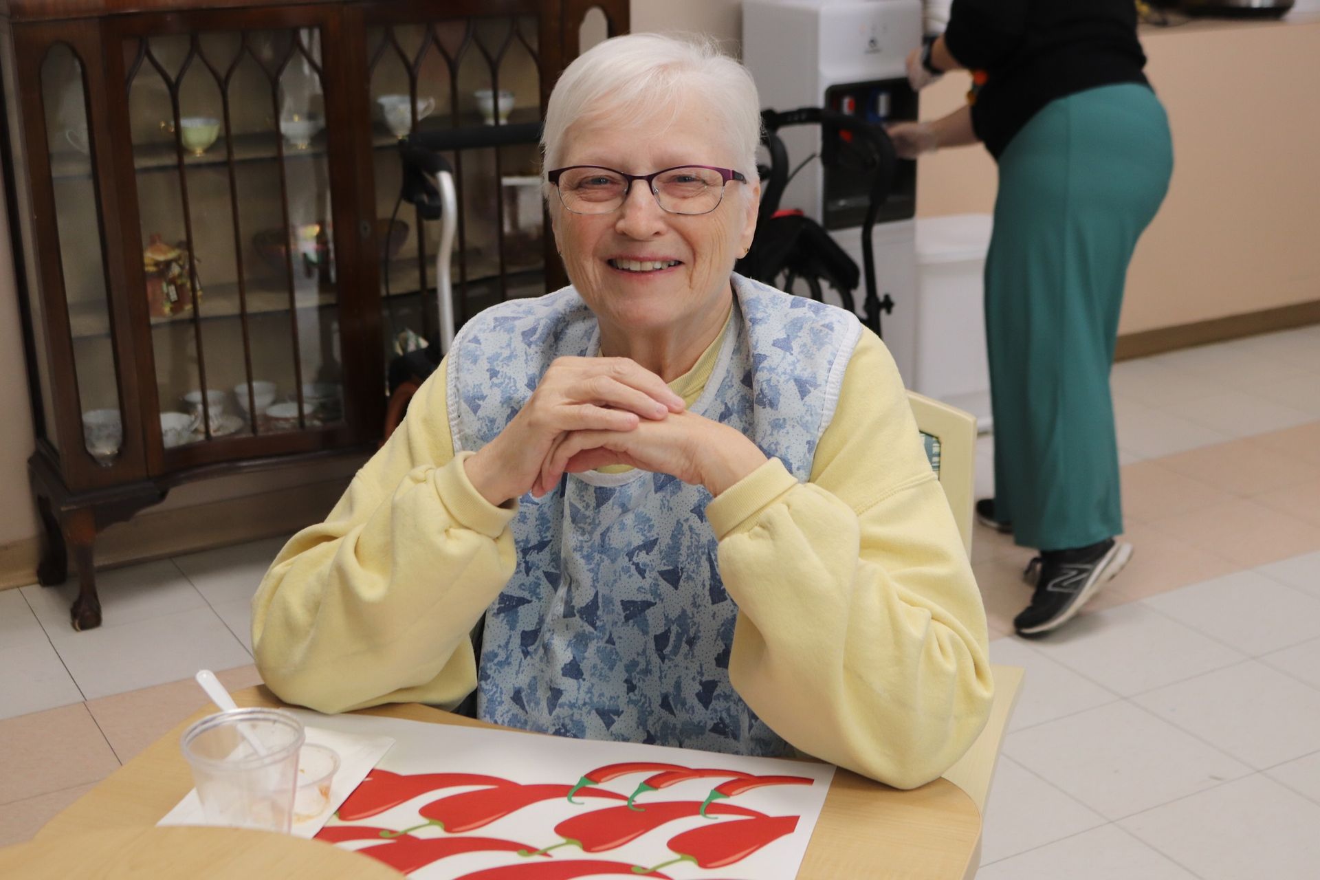 Smiling elderly woman seated at a table with a red-and-white placemat in a bright indoor setting