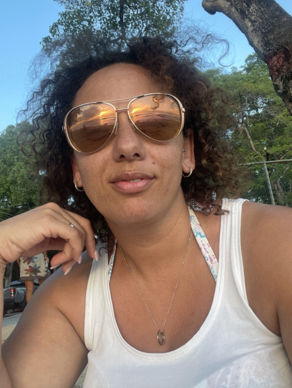 Woman wearing sunglasses and a white tank top, sitting outdoors with trees in the background.