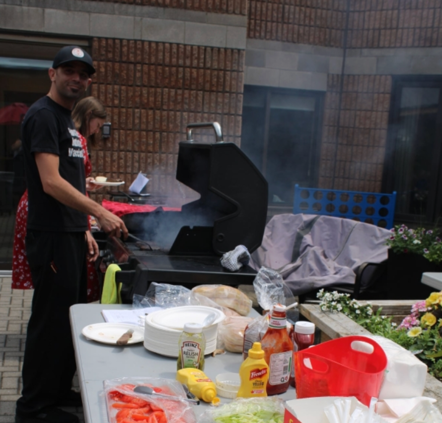Person grilling outdoors beside a picnic table with food, drinks, and red containers