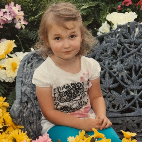 Smiling child seated on a gray bench among colorful flowers in a garden setting