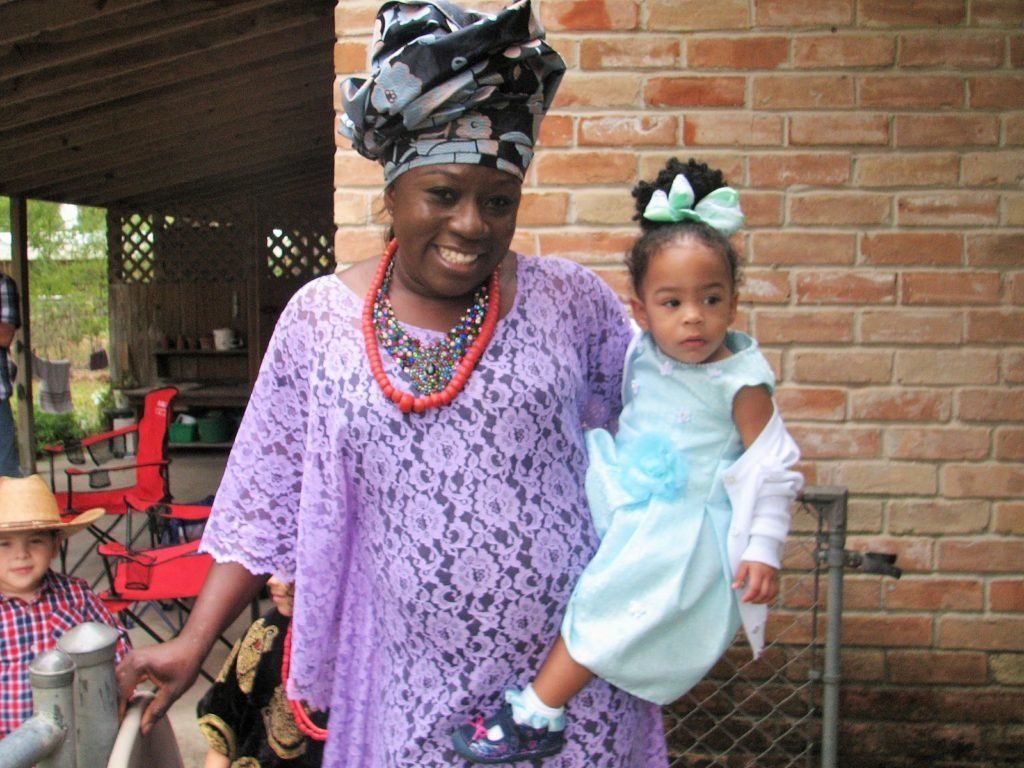 Woman in purple lace dress holds a young child in a blue dress. They are outside near a brick wall.