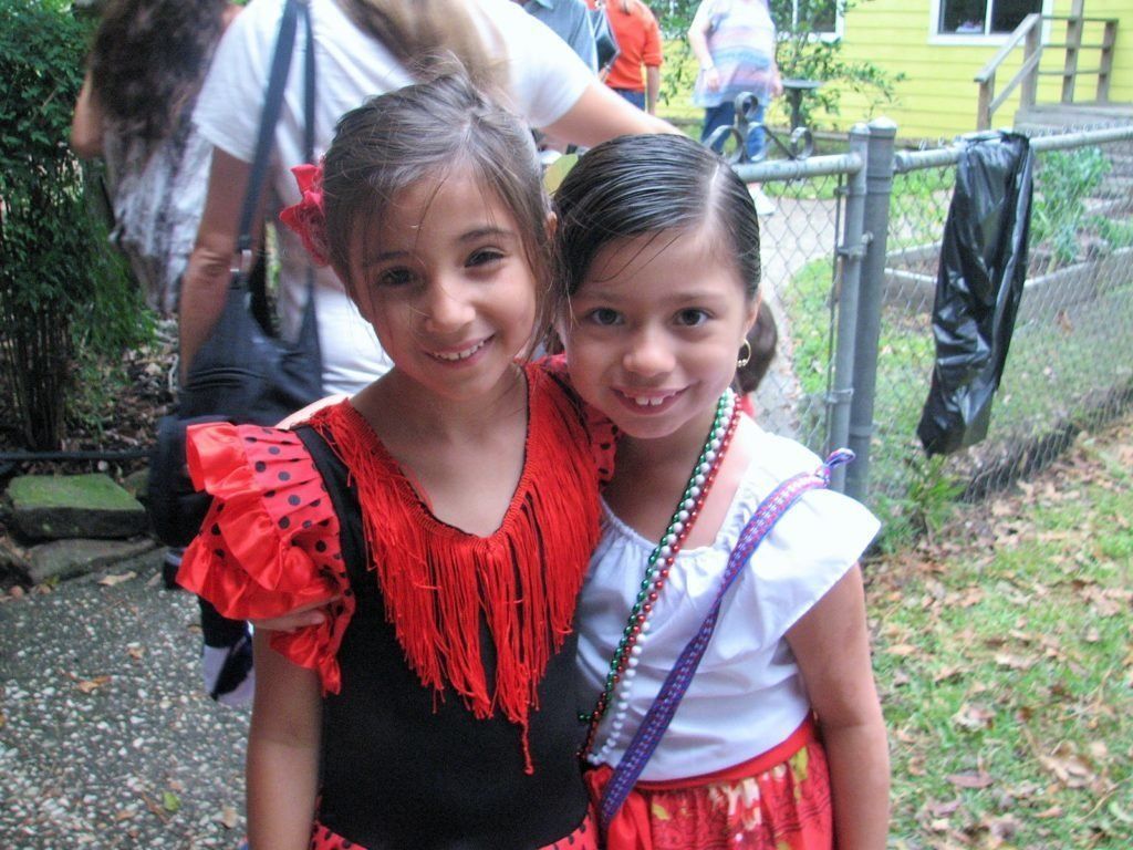 Two Montessori childs, one in red fringed top, the other in white, smiling and posing together outdoors.