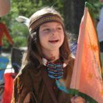 Montessori Child in Native American-style costume, smiling, holding flag, outdoor setting.