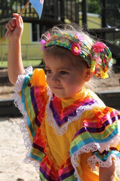 Montessori child in colorful dress, headband, and holding a flag, possibly at a festival outdoors.
