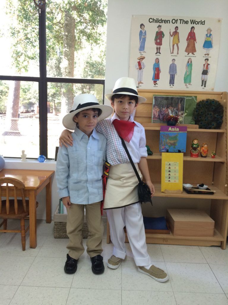 Two Montessori boys in costumes; one has arm around the other's shoulder. They wear hats, shirts, pants, and a shoulder bag.