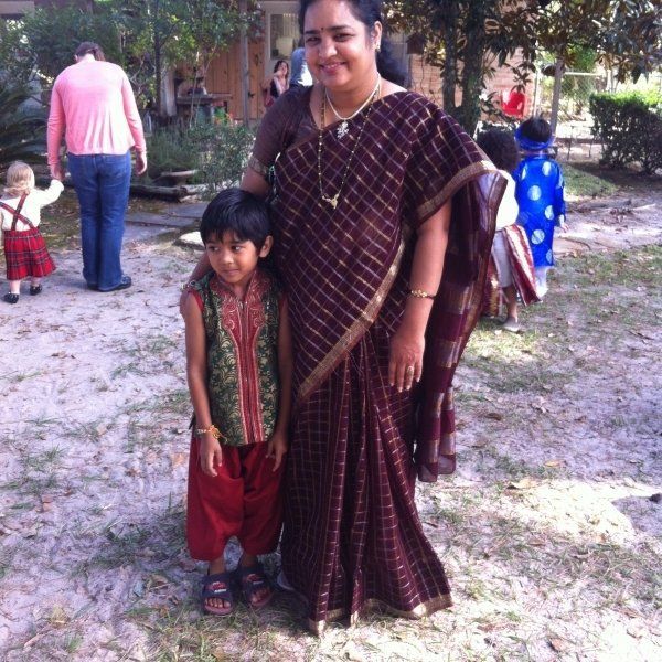 Woman in maroon saree with a child in traditional attire standing outside; other people in the background.