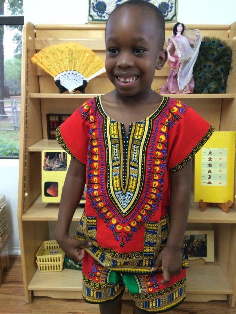 Young child wearing a red and yellow dashiki outfit smiles, standing in front of a bookshelf.