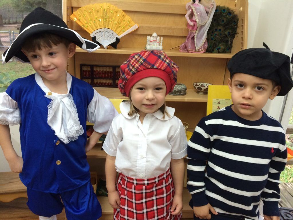 Three Montessori children in costume: one in blue, one in Scottish attire, one in a striped shirt and beret, standing in front of a bookshelf.